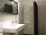 Moderne badeværelse med rektangulær vask, væghængt vandhane, stort spejl og en brusekabine med hvide, kvadratiske fliser og et sort bruseforhæng.