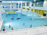 Indendørs swimmingpool med flere mennesker, der svømmer og leger i vandet; moderne design, hvide flisegulve og gule stole ses i baggrunden.