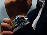 En person iført et sølvfarvet armbåndsur med sort urskive og blå-rød ramme, klædt i en mørk jakke og lys sweater, justerer uret på sit venstre håndled.