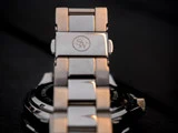 Nærbillede af et metalarmbånd med S/W-logoet indgraveret på låsen, og en del af urkassen er synlig i baggrunden.
