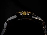 Nærbillede af en armbåndsurskrone med gulddetaljer og et indgraveret logo mod en sort baggrund, der fremhæver oversiden af uret.