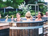En gruppe mennesker slapper af i et udendørs boblebad, smilende og med champagneglas i hånden, omgivet af grønne områder og et træhegn.
