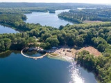 Luftfoto af en sø omgivet af tæt, grøn skov med en cirkulær træbrygge og et byggeområde langs bredden i sollys.