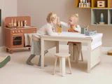 To små børn leger og tegner ved et hvidt bord i et pænt, pastelfarvet legeværelse med et legetøjskøkken og hylder i baggrunden.