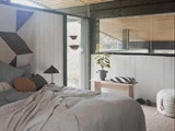 Et hyggeligt soveværelse med en uredt seng, en sort lampe, en træbænk med en potteplante og en stribet pude, store vinduer og blødt naturligt lys.