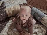En baby iført en brun trøje og grå sokker sidder på et mønstret tæppe, omgivet af en blød, flerfarvet pude.