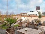 En tagterrasse med træborde og -stole, potteplanter og byens bygninger i baggrunden under en delvis overskyet himmel.