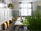 En lys og hyggelig café med hvide borde, bænke med hynder, gule puder, planter og sollys, der strømmer ind gennem et stort vindue med tynde gardiner.