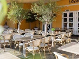 Udendørs restaurantterrasse med træborde, hvide stole og pottetræer mod en gul væg og hvide dobbeltdøre.