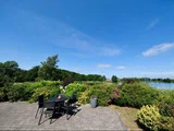 En terrasse med et sort bord og stole har udsigt til en frodig have, en sø og træer under en klar blå himmel.