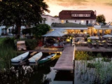 Udendørs spiseplads ved solnedgang med folk siddende under parasoller ved en kaj, tre fortøjede både, frodigt grønt og et stort hus i baggrunden.