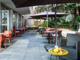 Udendørs gårdhave ved en café med forskellige stole og borde, skygget af store parasoller, omgivet af planter og grønt. Drikkevarer og kager står på et gult bord i forgrunden.