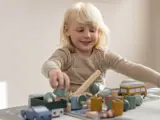 Et lille barn med lyst hår leger ved et bord med legetøjsbiler, træer og bygninger af træ, mens det smiler og fokuserer på aktiviteten.
