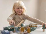 Et lille barn med lyst hår smiler, mens det leger med legetøjsbiler, træer og bygninger af træ på et bord.