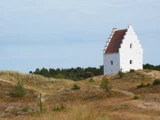 En lille hvid kirke med aftrappet gavltag står alene på græsklædte klitter, omgivet af sparsomme træer under en lyseblå himmel.