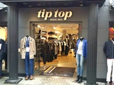 Indgangen til en tøjbutik med sorte søjler, et skilt med teksten »tip top« over døren og to mannequiner på hver side, klædt i henholdsvis festligt og afslappet tøj. Inde i butikken kan man se stativer
