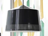 En sort metalpendel med lodrette slidser nær den nederste kant, der hænger foran et vindue med farverige, geometrisk mønstrede gardiner.