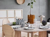 Et rundt spisebord i træ med stribet service, glas, en potteplante, grønt glas, vindruer og en lampe, omgivet af lyse træstole i et moderne, minimalistisk rum.