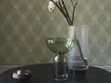 En grøn glasvase, en klar ribbet vase med blomstrende grene og to små fade står på et mørkt bord mod et mønstret grønt tapet.