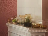 Glasvaser og skåle med grønne planter er arrangeret på en hvid kamin mod et rosa blomstertapet og en hvid flisevæg.