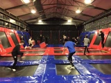 Flere mennesker hopper og leger på sammenkoblede trampoliner i en indendørs trampolinpark med rød, sort og blå polstring.