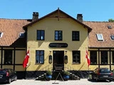 En gul bygning med rødt tegltag, to danske flag ved indgangen og et skilt med teksten Tranekær Slotskro. To biler holder parkeret foran under en klar blå himmel.