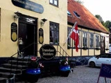 En gul bygning med skilte, hvor der står Tranekaer Slotskro, dekoreret med danske flag, udendørs trapper og en parkeret hvid bil ved siden af.