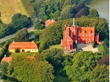 Luftfoto af et stort slot af røde mursten med tårne, omgivet af træer, en dam og nærliggende bygninger med gule tage i et frodigt grønt landskab.