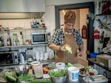 En kvinde i farverigt tøj og forklæde tilbereder mad i et køkken, mens hun presser sauce ud på et fad omgivet af friske ingredienser og køkkenredskaber.
