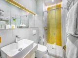 Moderne badeværelse med hvidt inventar, håndvask, toilet, stort spejl og en glasbruser med guldmosaikfliser på bagvæggen. Håndklæder hænger på højre væg.
