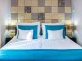 En pænt redt hotelseng med hvidt sengetøj, blågrønne detaljer og firkantede pyntepuder mod et polstret geometrisk hovedgærde. Der står lamper på hver side af sengen.