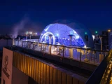 En bar på taget om aftenen har en glødende geodætisk kuppel oplyst med blå lys, udendørs siddepladser og lyskæder langs rækværket med udsigt over byens skyline.