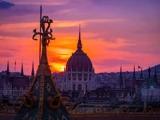 En levende solnedgangshimmel bag den kuppelformede ungarske parlamentsbygning i Budapest med udsmykkede hustage i silhuet i forgrunden.