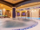 Indendørs spa-område med et rundt spabad i forgrunden, liggestole ved poolen til venstre og en swimmingpool i baggrunden med en varm, stemningsfuld belysning.
