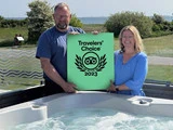 To smilende mennesker står ved et spabad og holder et grønt Travelers' Choice 2023-skilt udendørs med grønne planter og vand i baggrunden.