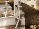 En sæbedispenser i klart glas med en sølvfarvet pumpe står på en træplade ved siden af en quiltet brun pude og et spejl.