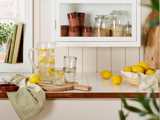 Et køkkenbord med en kande citronvand, to glas, hele citroner, en kurv med citroner, et skærebræt og krukker med tørvarer i et skab ovenover.