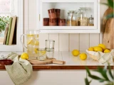 Et køkkenbord med en kande citronvand, to glas, hele citroner, en kurv med citroner, et skærebræt og krukker med tørvarer i et skab ovenover.