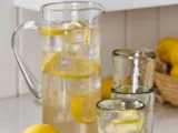 En glaskande med isvand og citronskiver ved siden af tre tomme glas og friske citroner i en kurv på køkkenbordet.