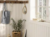 Hyggeligt hjørne med en potteplante, bøger, hækletaske, vaser med blomster, trækroge og sollys, der strømmer ind gennem et vindue på vægge med cremefarvet panel.