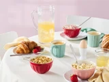Morgenbord med kornskåle, croissanter, et blødkogt æg, jordbær, krus med kaffe, et glas og en kande med juice, alt sammen dækket på en hvid dug.