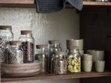 Glaskrukker med korklåg fyldt med bønner, kerner og marineret ost står på en træhylde ved siden af beige keramiske kopper og skåle.