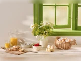 Et morgenbord med brød, jordbær, en kande appelsinjuice, en skål med æg, tallerkener og en vase med blomster ved siden af et grønt vindue.