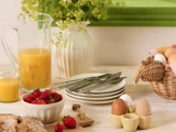 Et morgenbord med jordbær, æg, appelsinjuice, tallerkener, bestik, brød og en vase med blomster på en lys træoverflade.