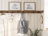 En cremefarvet skjorte og en grå hæklet taske hænger på trækroge under indrammede skibsskitser; en vase med blomster, et håndspejl og grønne planter pynter på scenen.