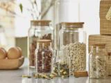 Glaskrukker fyldt med forskellige tørrede bønner og frø står på køkkenbordet ved siden af en træskål med æg og flettede kurve i baggrunden.