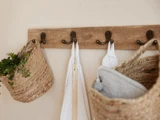 Et vægstativ i træ med fire metalkroge, der holder flettede kurve med planter og håndklæder, som hænger på krogene.