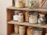 Glaskrukker fyldt med korn, nødder og krydderier er pænt arrangeret på træhylder med stablede beige kopper på den nederste hylde. Opstillingen ser ryddelig og organiseret ud.