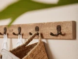 Et nærbillede af en knagerække i træ med fire metalkroge, der holder en vævet taske og hvidt stof mod en beige væg. Et grønt blad dækker delvist det øverste venstre hjørne.