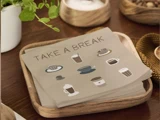 En stak servietter med "TAKE A BREAK" og illustrationer af forskellige kaffekopper, placeret på en træbakke på et bord med planter og køkkenting.